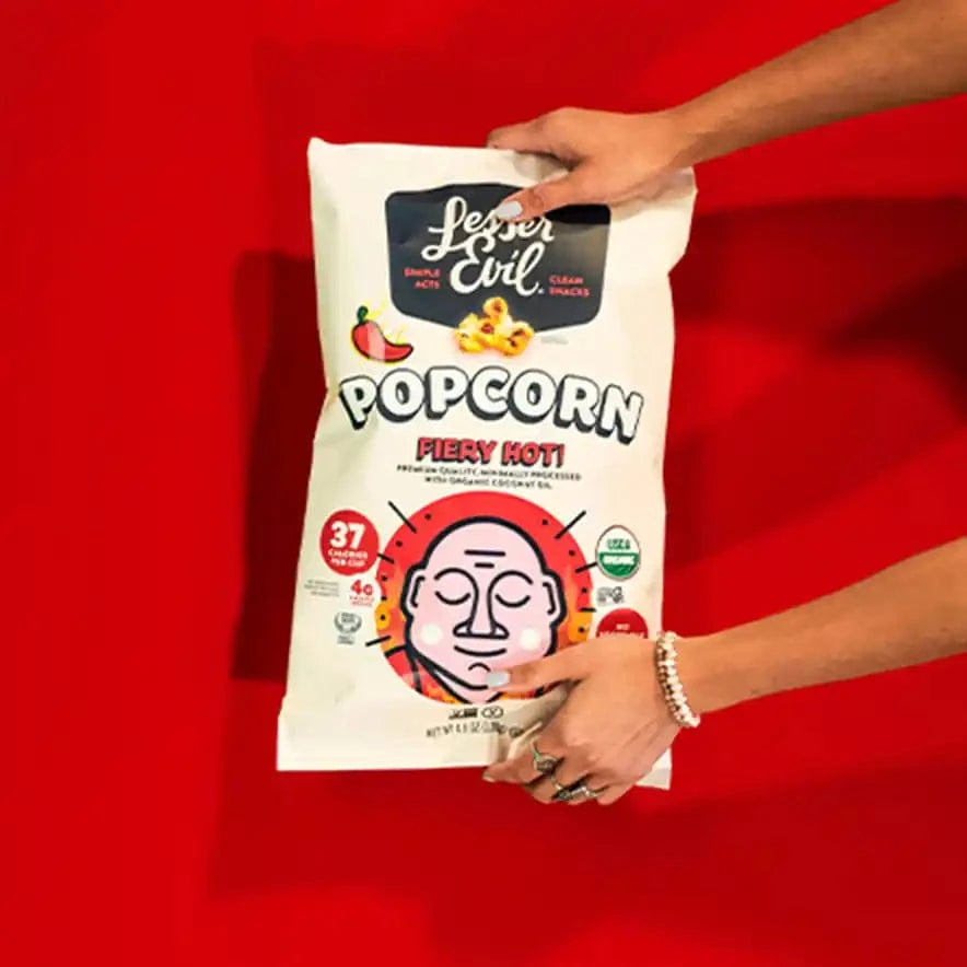 Two hands hold a Big Flavor Popcorn Box by LesserEvil Snacks against a bright red background. The box shows a cartoon face and spicy pepper illustrations.