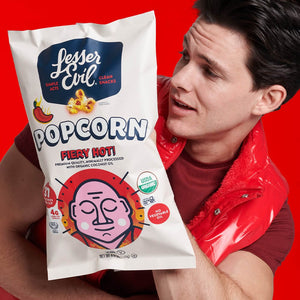 A person in a red vest holds a large bag of LesserEvil Fiery Hot Organic Popcorn against a bright red background, looking at the bag with interest.