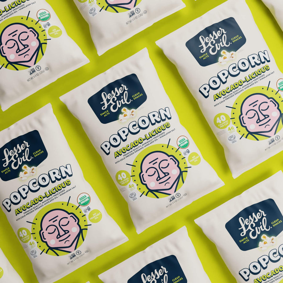 LesserEvil Avocado-licious Organic Popcorn bags are arranged on a bright green background, with packaging showing a stylized pink face and green accents.