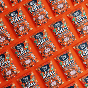 Several packets of LesserEvil Sweet Potato Apple Asteroid Lil Puffs feature bright orange packaging, playful cartoons, and proudly display their 0g added sugar in a repeating pattern.