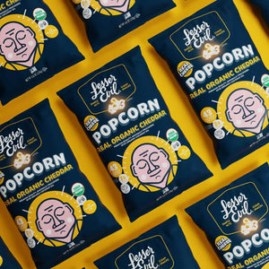 Bags of OrganicPopcorn Real Organic Cheddar Popcorn, with cartoon faces and bold popcorn labels, are displayed on a yellow background.