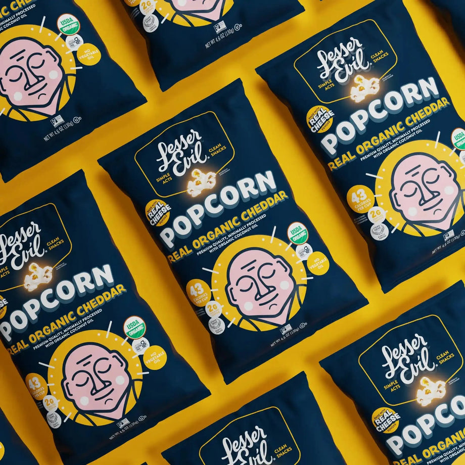 Bags of OrganicPopcorn Real Organic Cheddar Popcorn, with cartoon faces and bold popcorn labels, are displayed on a yellow background.