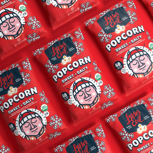LesserEvil Sweet + Salty Popcorn in red, snowflake-adorned bags with a winter-hat cartoon—festive holiday snacks arranged diagonally.