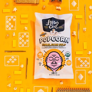 A Popcorn Best Sellers Box from LesserEvil Snacks on a bright yellow background, surrounded by popcorn pieces, playing cards, golden pens, and small notebooks.