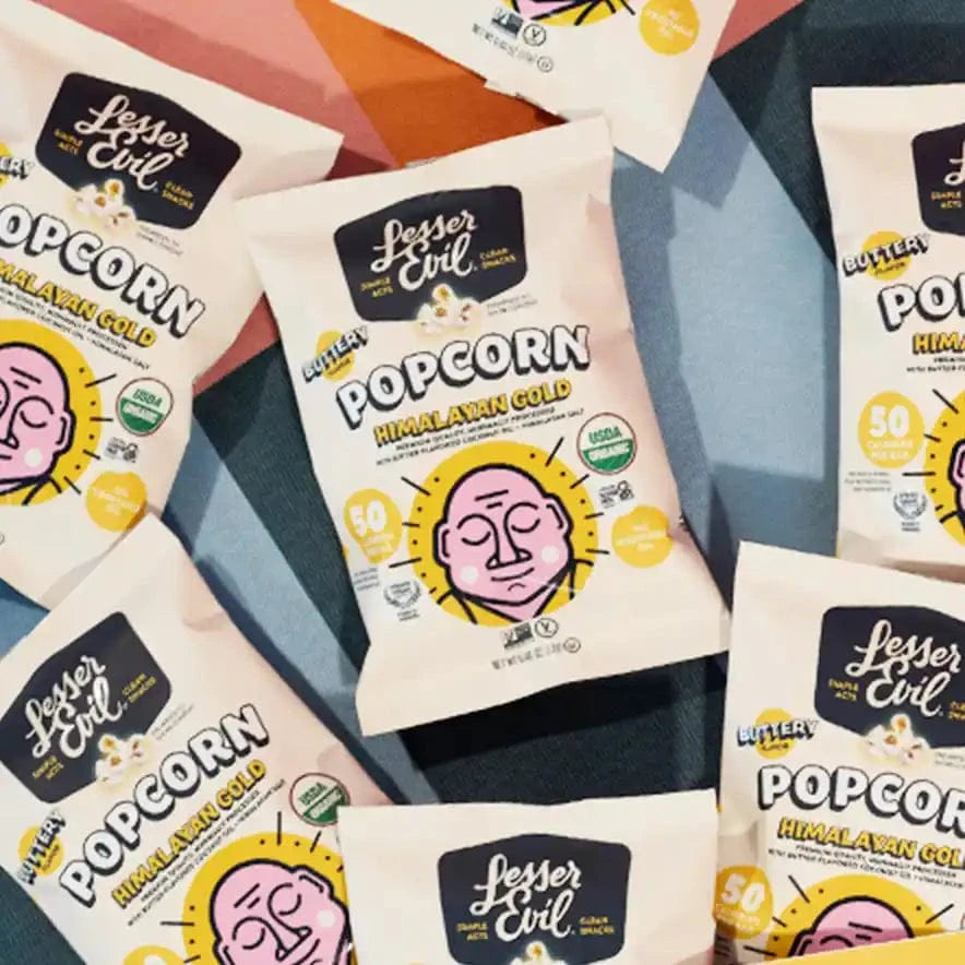 Himalayan Gold Organic Popcorn – 24 Pack by LesserEvil Snacks is displayed on a colorful geometric background with bold black and yellow text and a pink-faced cartoon.
