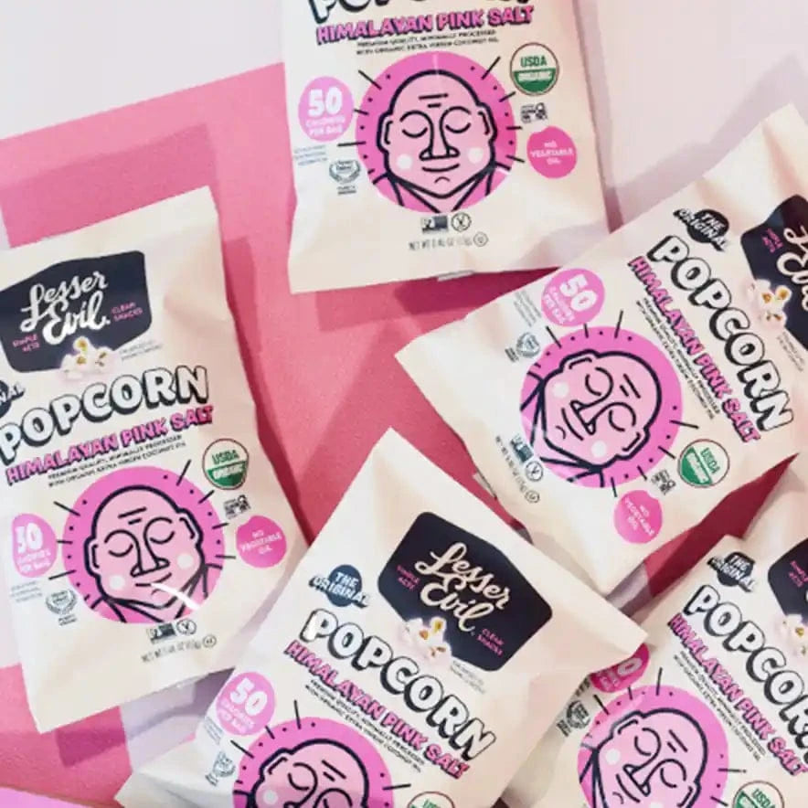 Several bags from the LesserEvil Snacks Popcorn Best Sellers Box are scattered on a pink and white surface, showing the brand’s logo and details.