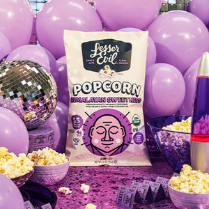 A box of The Classics Popcorn by LesserEvil Snacks is surrounded by purple balloons, a disco ball, a lava lamp, bowls of popcorn, and purple movie tickets.
