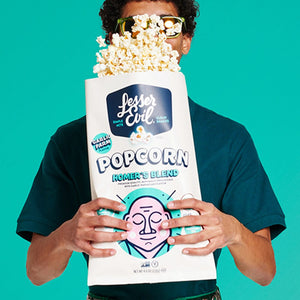 A person in sunglasses holds a giant bag of OrganicPopcorn’s Homers Blend Organic Popcorn with kernels spilling out, set against a teal background.