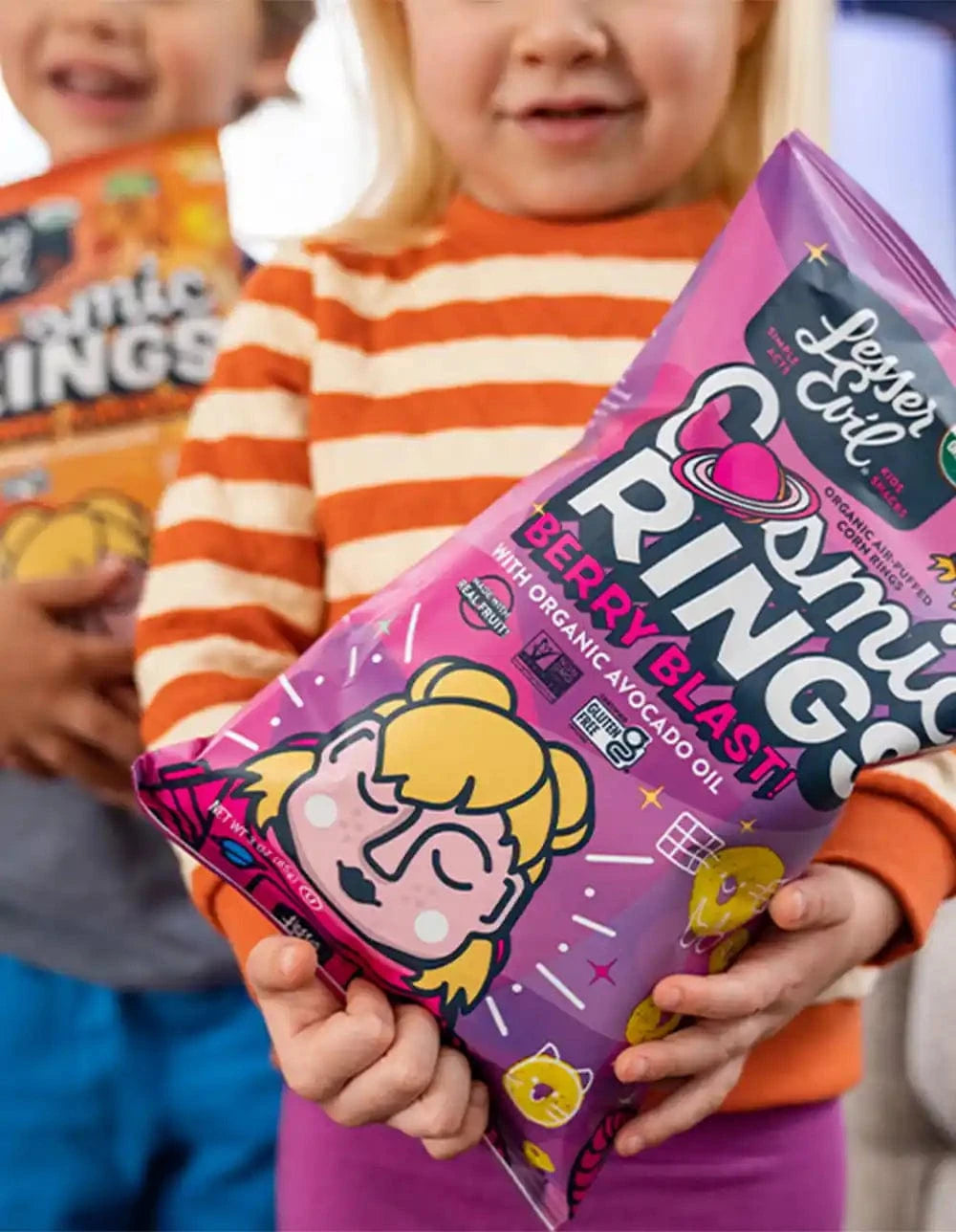 Two children holding snacks; one holds a purple bag of Lesser Evil Cosmic Rings Berry Blast, and the other holds an orange bag of the same brand.