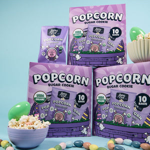 Three purple LesserEvil Snacks Easter Sugar Cookie Popcorn Snack Boxes and a small bag are shown with candy eggs and pastel decor on a blue background.