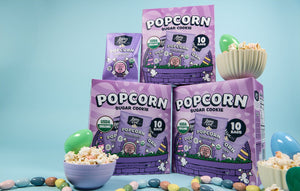 Three purple LesserEvil Snacks Easter Sugar Cookie Popcorn Snack Boxes and a small bag are shown with candy eggs and pastel decor on a blue background.