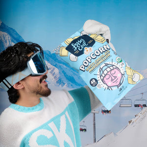 A man in ski gear holds a bag of LesserEvil White Chocolate Marshmallow popcorn, with snowy mountains and ski lifts in the background.