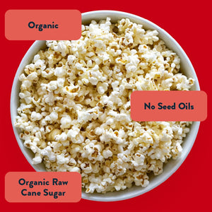 A bowl of LesserEvil Sweet + Salty Popcorn sits on a red background, labeled “Organic,” “No Seed Oils,” and “Organic Raw Cane Sugar”—a perfect holiday snack.