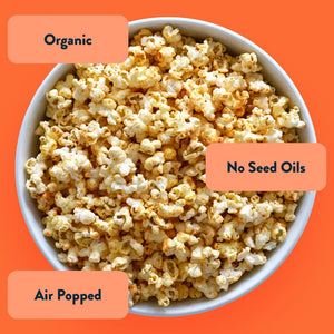 A bowl of Primal Kitchen Buffalo Popcorn by LesserEvil Snacks on an orange background, showcasing clean ingredients, organic, no seed oils, and air popped labels.