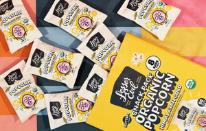 Eight small bags of LesserEvil Himalayan Gold Organic Butter Flavor Popcorn Snack Pack sit on a colorful striped surface beside a large snack box.