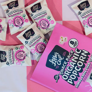 A pink and white checkered surface with snack-sized bags and a large pack of OrganicPopcorn’s Himalayan Pink Salt Popcorn Snack Pack—a tasty, clean snack in pretty packaging.