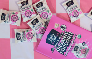 A pink and white checkered surface with snack-sized bags and a large pack of OrganicPopcorn’s Himalayan Pink Salt Popcorn Snack Pack—a tasty, clean snack in pretty packaging.