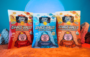 Three colorful bags of SpaceBalls Buffalo Cheddar Space Balls snacks stand on sand with rocks and a glowing blue-pink background, highlighting their organic ingredients.
