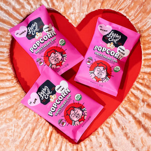 Three bright pink bags of Lesser Evil Sweetheart Pop popcorn are arranged on a heart-shaped red and peach background, featuring cartoon faces and heart-shaped labels.