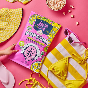 Flat lay of summer essentials on pink: yellow flip-flops, a hat, sunglasses, striped towel, bikini, popcorn, and LesserEvil Pink Lemonade Popcorn with lemon designs.