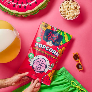 A person holds a Sweet Summer Box by LesserEvil Snacks, surrounded by green shorts, sunglasses, a beach ball, and popcorn on a pink surface—summer snacking ready!.