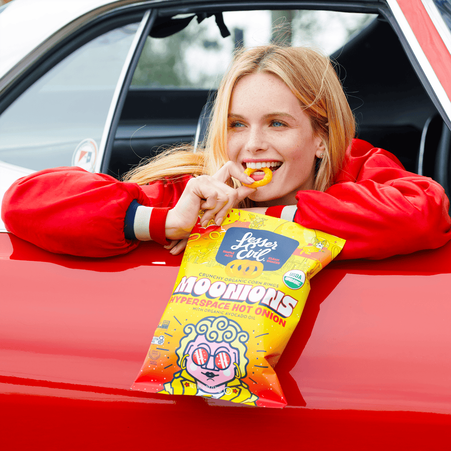 A smiling woman in a red jacket leans out of a red car, enjoying LesserEvil Hyperspace Hot Onion Moonions from a yellow bag.