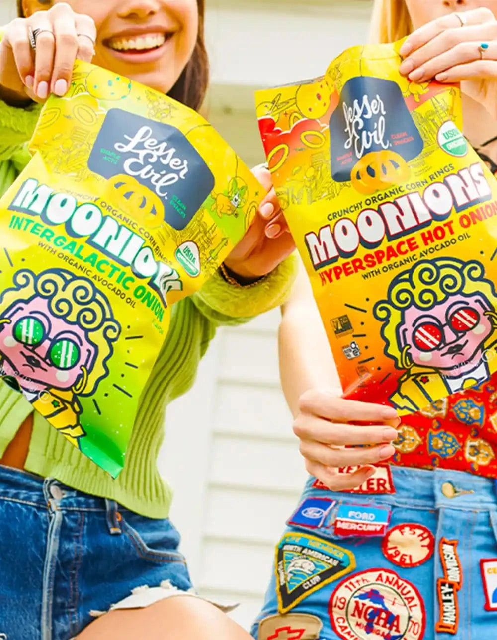 Two women holding colorful bags of Lesser Evil Moonions organic corn rings, showing Intergalactic Onion and Hyperspace Hot Onion flavors.