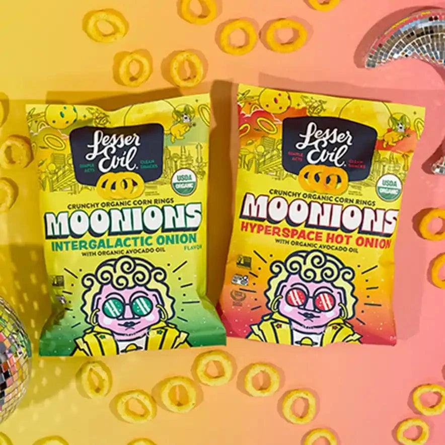 Two colorful boxes of Rheas Recos Moonions by LesserEvil Snacks sit on a pink and yellow surface, surrounded by crunchy corn rings and disco balls.