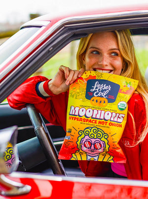 A smiling woman in a red jacket sits in a car, holding a bright yellow bag of LesserEvil Hyperspace Hot Onion Moonions made with zesty spices and avocado oil.