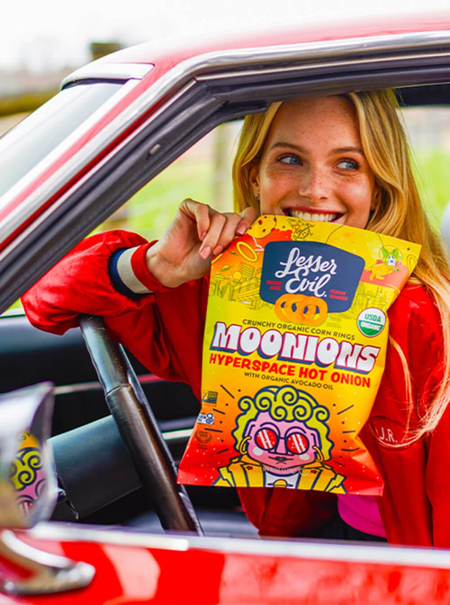 A smiling woman in a red jacket sits in a car, holding a bright yellow bag of LesserEvil Hyperspace Hot Onion Moonions made with zesty spices and avocado oil.