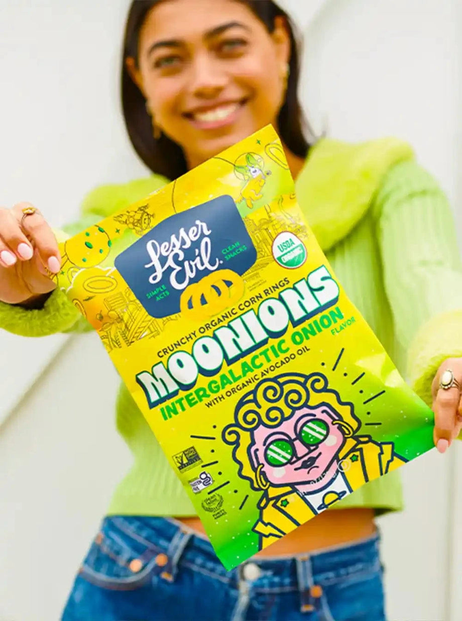 A smiling woman in a green sweater holds a box of LesserEvil Snacks Snacktime Box close to the camera.