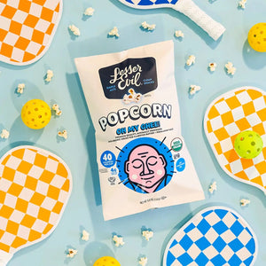 A Dairy Good Snacks Box by LesserEvil Snacks is surrounded by orange and blue pickleball paddles, yellow balls, and scattered popcorn on a light blue background.