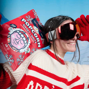 A smiling person in ski goggles and red gloves holds a bag of LesserEvil Sweet + Salty Popcorn, the perfect holiday snack, wearing a festive sweater with fake snow on their head.