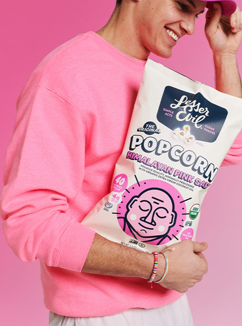 A smiling person in a pink sweatshirt holds a bag of OrganicPopcorn’s Himalayan Pink Salt Organic Popcorn against a bright pink background.