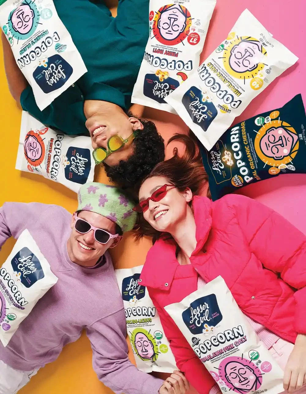 Two smiling people in colorful outfits and sunglasses lie on a pink and orange background, surrounded by bags of Lesser Evil popcorn in different flavors.
