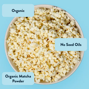 A bowl of LesserEvil White Chocolate Marshmallow popcorn on a blue background with labels: Organic, No Seed Oils, Organic Matcha Powder—a perfect holiday sweet snack.