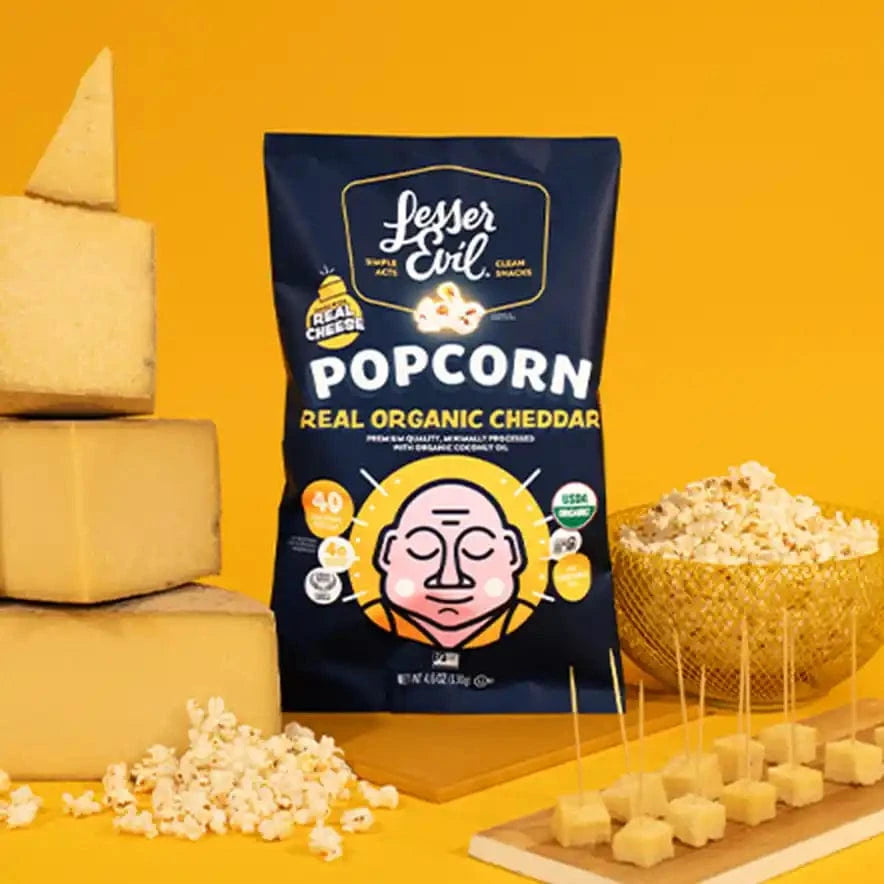 A Popcorn Best Sellers Box from LesserEvil Snacks is displayed with cheddar cheese blocks, cheese cubes on toothpicks, and a bowl of popcorn on a yellow background.