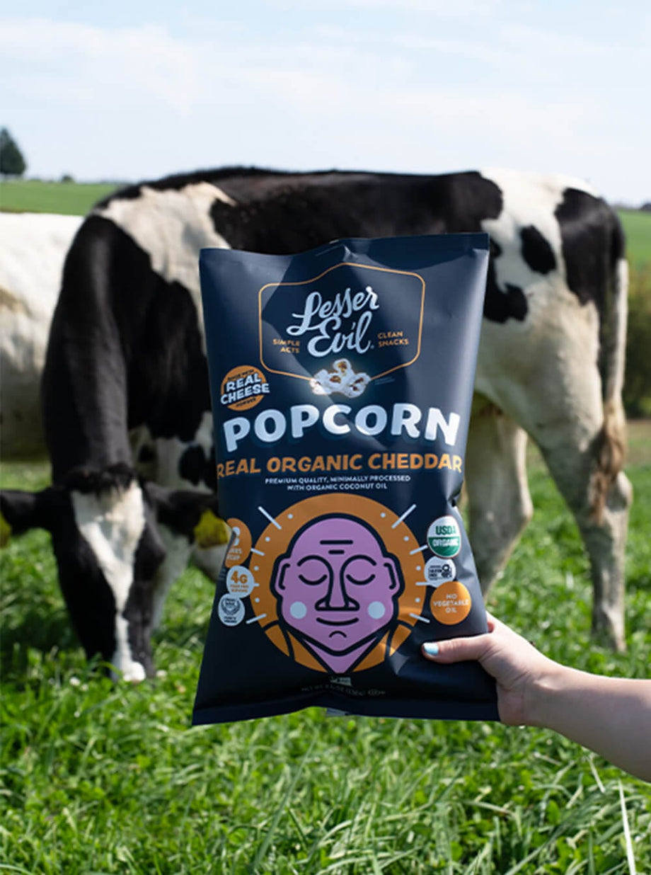 A hand holds a bag of OrganicPopcorn Real Organic Cheddar Popcorn in front of grazing cows in a grassy field.