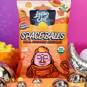 A bag of SpaceBalls Real Organic Cheddar Space Balls stands upright among cheese balls and silver spheres on a vibrant pink and orange background.