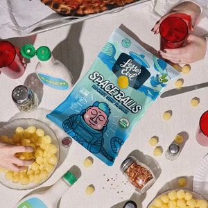 A table with pizza, red drinks, SpaceBalls Rocket Ship Ranch bottles, a bag of Rocket Ship Ranch Space Balls, cheese balls, and hands reaching in.