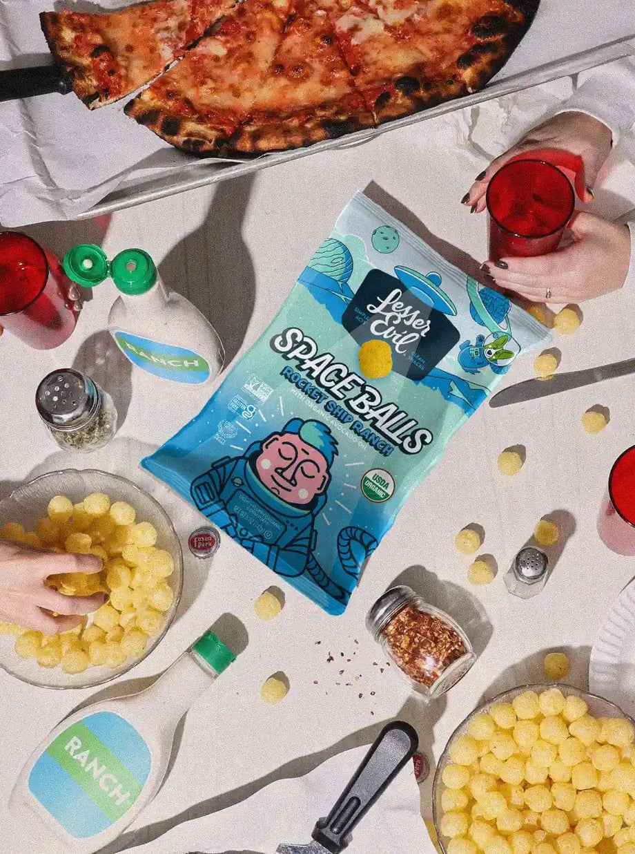A table with pizza, red drinks, SpaceBalls Rocket Ship Ranch bottles, a bag of Rocket Ship Ranch Space Balls, cheese balls, and hands reaching in.