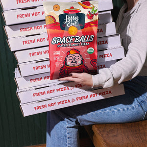 A woman sits on a wooden chair holding pizza boxes and a bag of SpaceBalls Outer Planet Pizza Space Balls with dairy-free cheese.