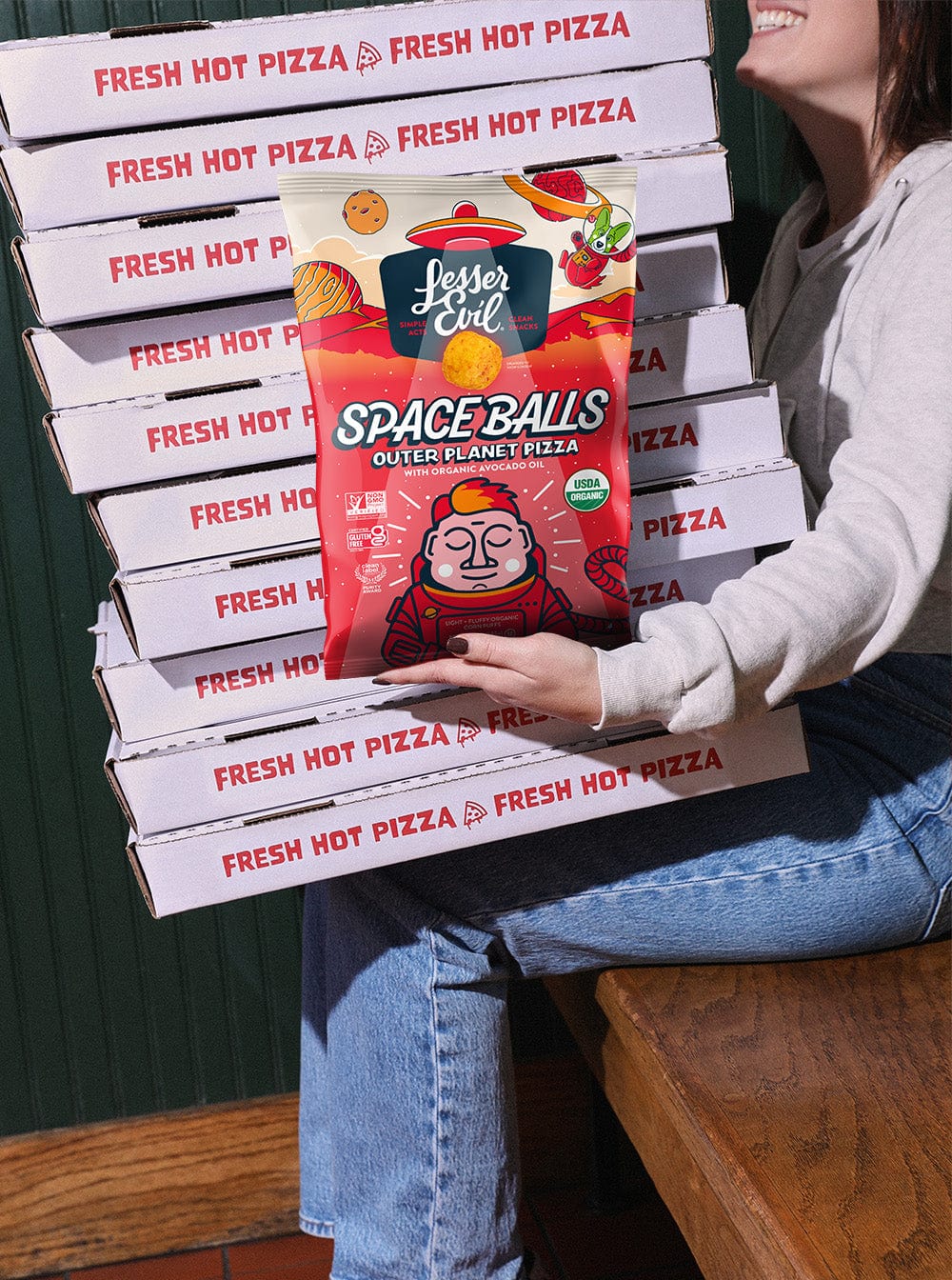 A woman sits on a wooden chair holding pizza boxes and a bag of SpaceBalls Outer Planet Pizza Space Balls with dairy-free cheese.