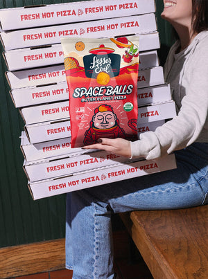 A woman sits on a wooden chair holding pizza boxes and a bag of SpaceBalls Outer Planet Pizza Space Balls with dairy-free cheese.