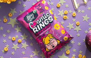 A pink bag of LesserEvil Berry Blast Cosmic Rings cereal, made with organic fruit powders, rests on a purple surface with scattered rings and glow-in-the-dark stars.