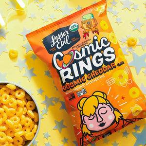 A bag of LesserEvil Cosmic Cheddar Cosmic Rings rests on a yellow surface with star confetti, beside a bowl of the ring-shaped snacks.