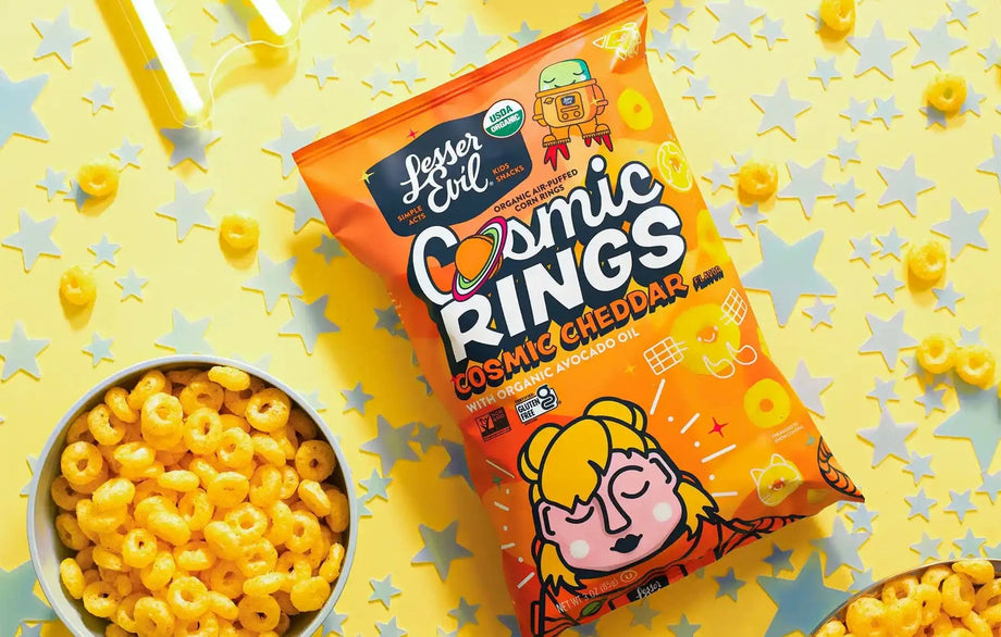 A bag of LesserEvil Cosmic Cheddar Cosmic Rings rests on a yellow surface with star confetti, beside a bowl of the ring-shaped snacks.