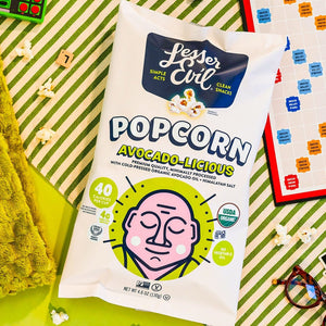 A bag of LesserEvil Avocado-licious Organic Popcorn rests on a green table with Scrabble tiles, glasses, a green towel, and popcorn pieces around it.