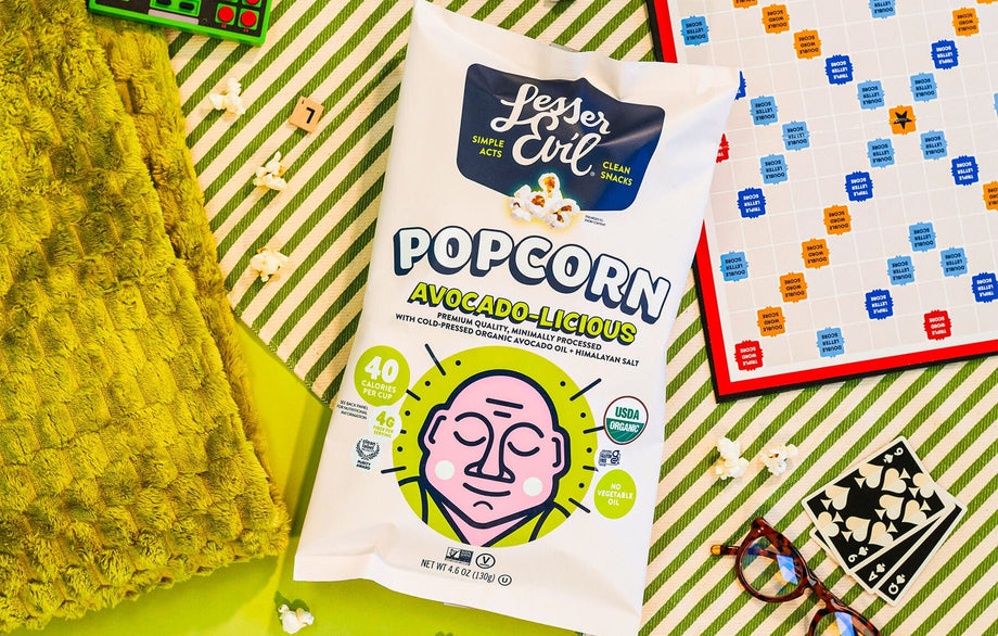 A bag of LesserEvil Avocado-licious Organic Popcorn rests on a green table with Scrabble tiles, glasses, a green towel, and popcorn pieces around it.