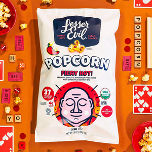 A bag of LesserEvil Fiery Hot Organic Popcorn sits on an orange surface surrounded by popcorn, game cards, Scrabble tiles, tickets, and small bowls.